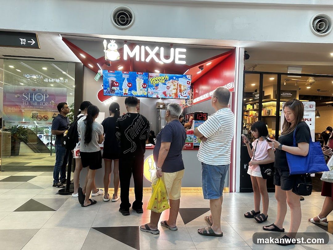 A Mixue store with a red and white facade. A line of customers is waiting to order. The menu board displays various drinks and ice cream.