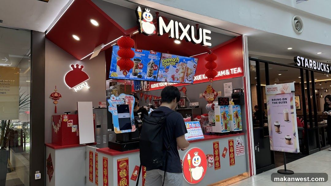 A Mixue Ice Cream & Tea shop front with a red and white color scheme. The shop features a large illuminated sign with the MIXUE logo. Digital menu boards display various drinks and prices. Red lanterns are hung as decorations. A customer is standing at the counter. Adjacent to the shop is a Starbucks.
