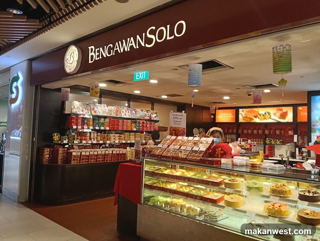 A bakery storefront with a display of cakes and pastries in refrigerated cases. Boxes of cookies and other baked goods are stacked on shelves behind the counter. A sign above the entrance reads "BENGAWAN SOLO". There are decorative lanterns hanging from the ceiling.