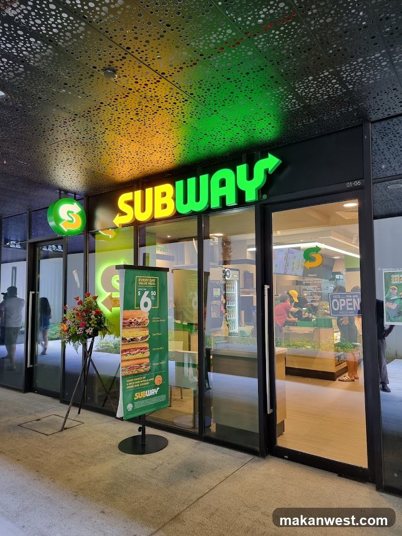 The exterior of a Subway restaurant with a prominent illuminated sign. A green banner displays sandwich options and a promotion. Inside, staff members are visible behind a counter.