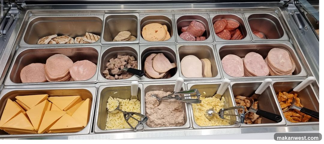 A refrigerated deli counter displays various food items in stainless steel trays. The top row contains grilled chicken strips, sliced processed meats, and sliced salami. The middle row features more sliced processed meats, diced meat, and sliced ham. The bottom row includes sliced cheese, shredded cheese, a creamy spread (possibly tuna or chicken salad), chopped egg salad, and pieces of cooked chicken. Scoops are present in several of the trays.