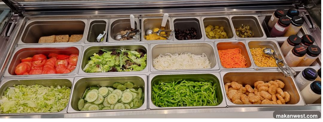 A refrigerated display case filled with various sandwich toppings and condiments. Visible toppings include sliced tomatoes, lettuce, sliced cucumbers, sliced green bell peppers, shredded carrots, onions, corn, olives, pickles, and what appear to be chicken nuggets. Several bottles of sauces and dressings are also present, including ketchup, mustard, barbecue sauce, chipotle southwest, spicy mayo, and sweet onion.