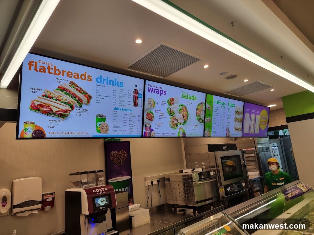 A digital menu board displaying various flatbreads, drinks, wraps, salads, subs, and toasties.