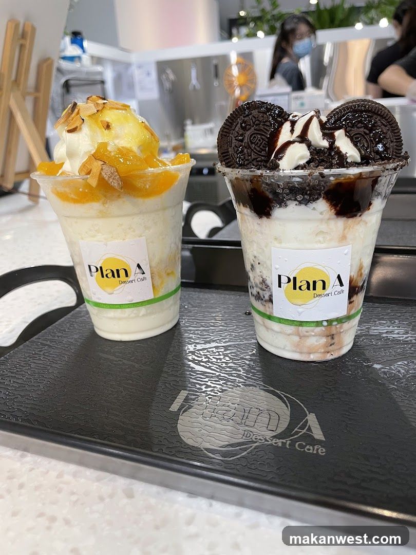 Two clear plastic cups filled with layered desserts sit on a dark gray tray. The cup on the left contains a white, fluffy base topped with yellow fruit, whipped cream, and slivered almonds. The cup on the right is layered with white cream, crushed dark cookies, and topped with whipped cream, chocolate syrup, and two whole cookies. Both cups have a sticker with the text "Plan A Dessert Cafe". The tray has a debossed logo that reads "Plan A Dessert Cafe". In the background, people are visible in a brightly lit cafe setting.