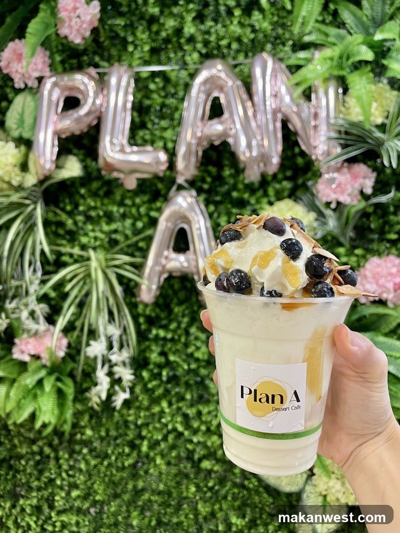A hand holds a clear plastic cup filled with a creamy, white dessert drink topped with whipped cream, blueberries, and slivered almonds. The cup has a label that reads "Plan A Dessert Cafe" and "Made from plants not oil." In the background, rose gold foil balloons spell out "PLAN A" against a backdrop of lush green artificial foliage and pink flowers.