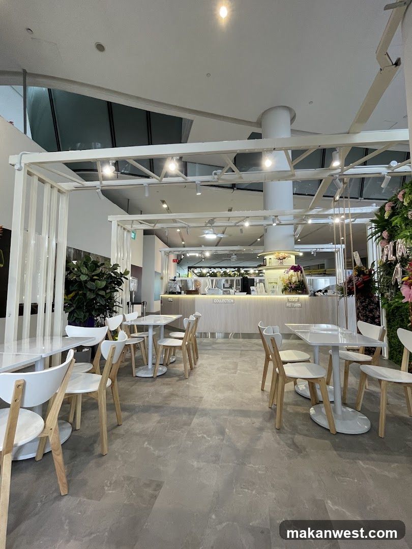 A bright and modern cafe interior with white tables and chairs arranged on a grey marble-patterned floor. The counter area features a light wood facade with signage. Plants are visible, adding a touch of greenery. The overall ambiance is clean and minimalist.