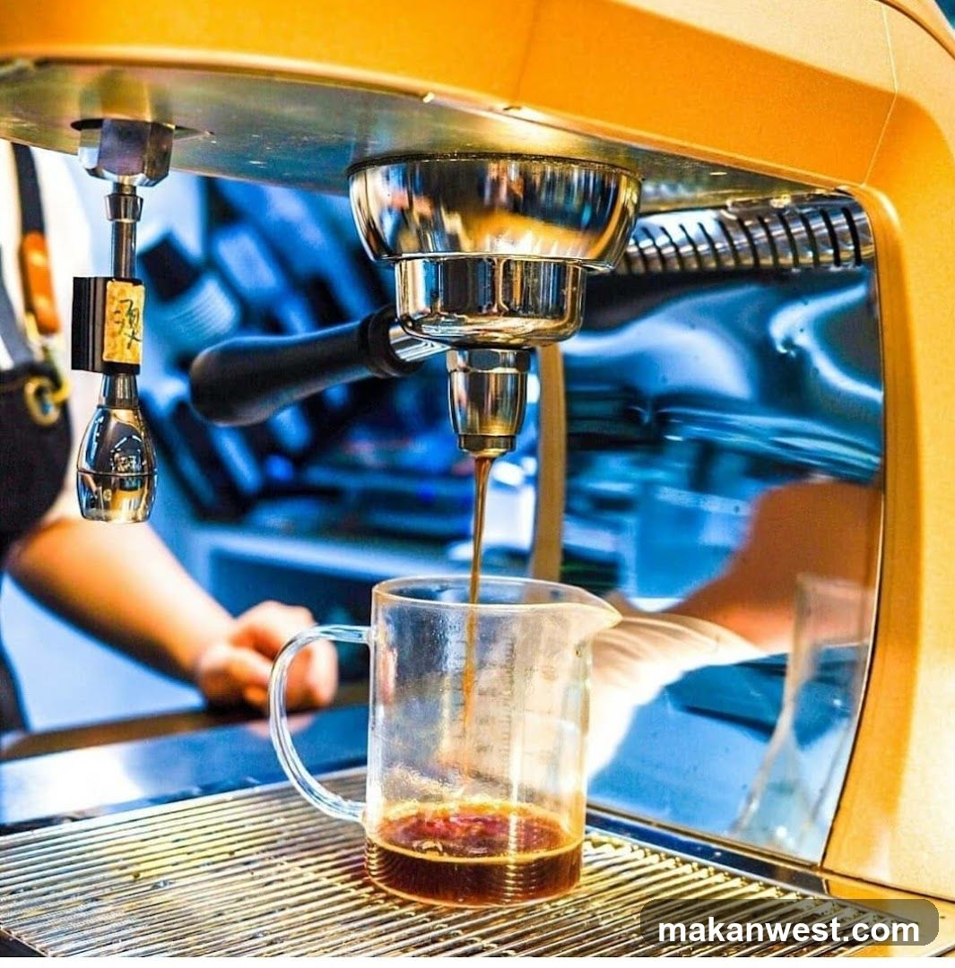 A close-up shot of an espresso machine brewing coffee into a glass pitcher. The machine is golden yellow and has a chrome portafilter. Coffee is seen dripping from the portafilter into the pitcher, which already contains a small amount of dark liquid. In the background, a person wearing an apron is visible, along with a blurred blue and white patterned surface. The overall scene suggests a coffee shop or cafe setting.