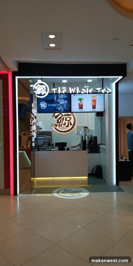 A modern bubble tea shop with a clean white counter and illuminated signage. The shop front features a prominent logo and digital screens displaying drinks. The interior is visible behind the counter, showing equipment for making beverages.