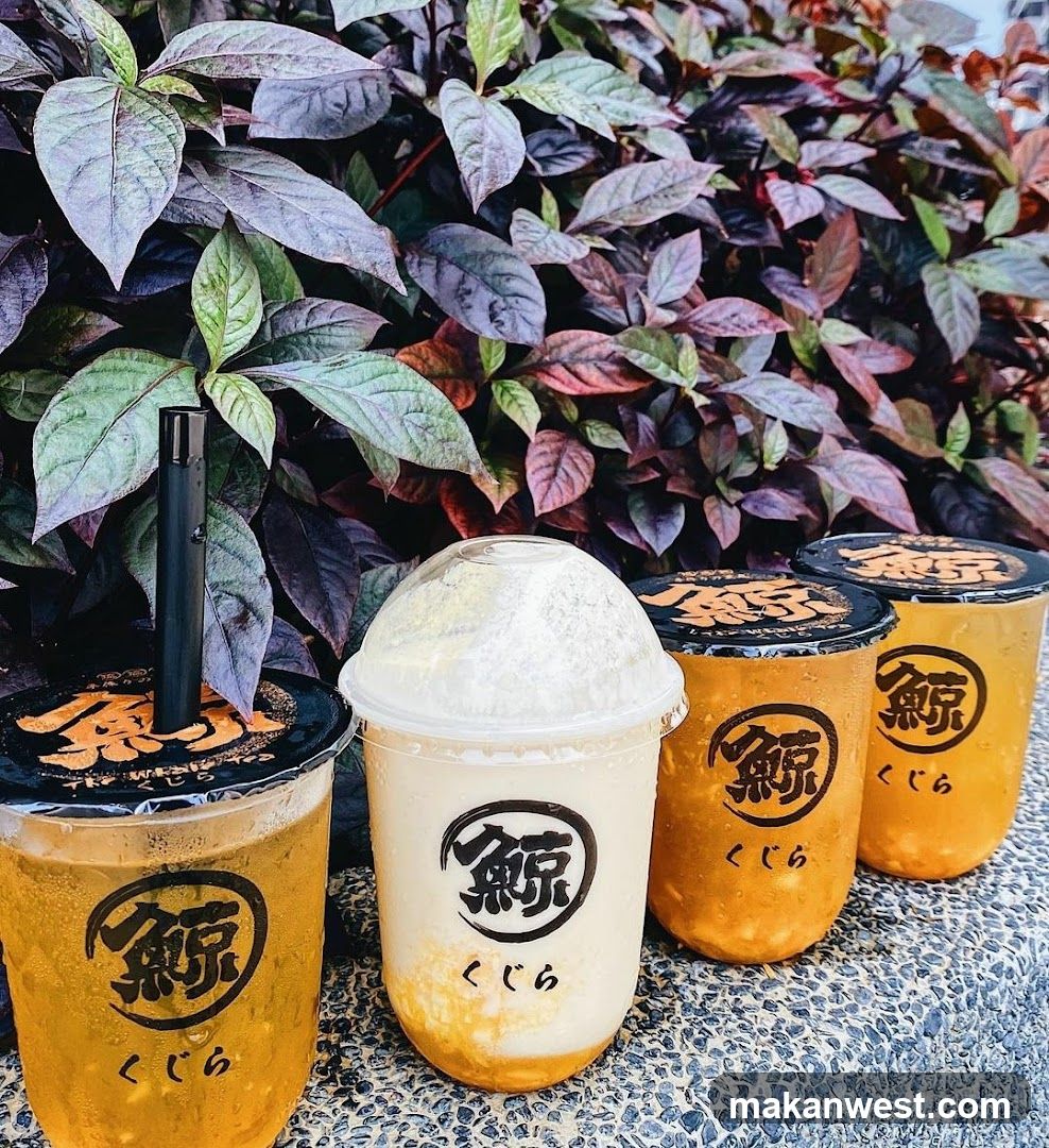 Four plastic cups with drinks are lined up in front of a dark purple and green leafy plant. The cups have black lids with an orange logo and Japanese characters. The first cup on the left has a black straw. The second cup from the left has a clear dome lid and appears to be a creamy beverage with a yellow layer at the bottom. The other two cups are filled with a golden-orange liquid.
