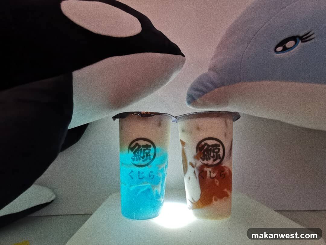 Two plastic cups with drinks are placed on a white surface. The cup on the left contains a blue liquid with white foam on top. The cup on the right contains a light brown liquid with white foam and some caramel-colored syrup. Both cups have a circular logo with Japanese characters and the word "くじら" (kujira) below it. A plush orca toy is on the left, and a plush dolphin toy is on the right, framing the drinks.