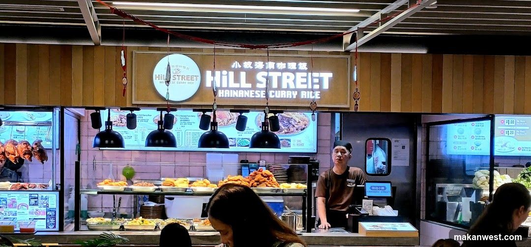 A food stall with a prominent sign reading "HILL STREET HAINANESE CURRY RICE". The stall displays various food items on shelves, including roasted meats hanging on the left. A staff member in a brown uniform stands behind the counter. Menu boards are visible above the food displays.