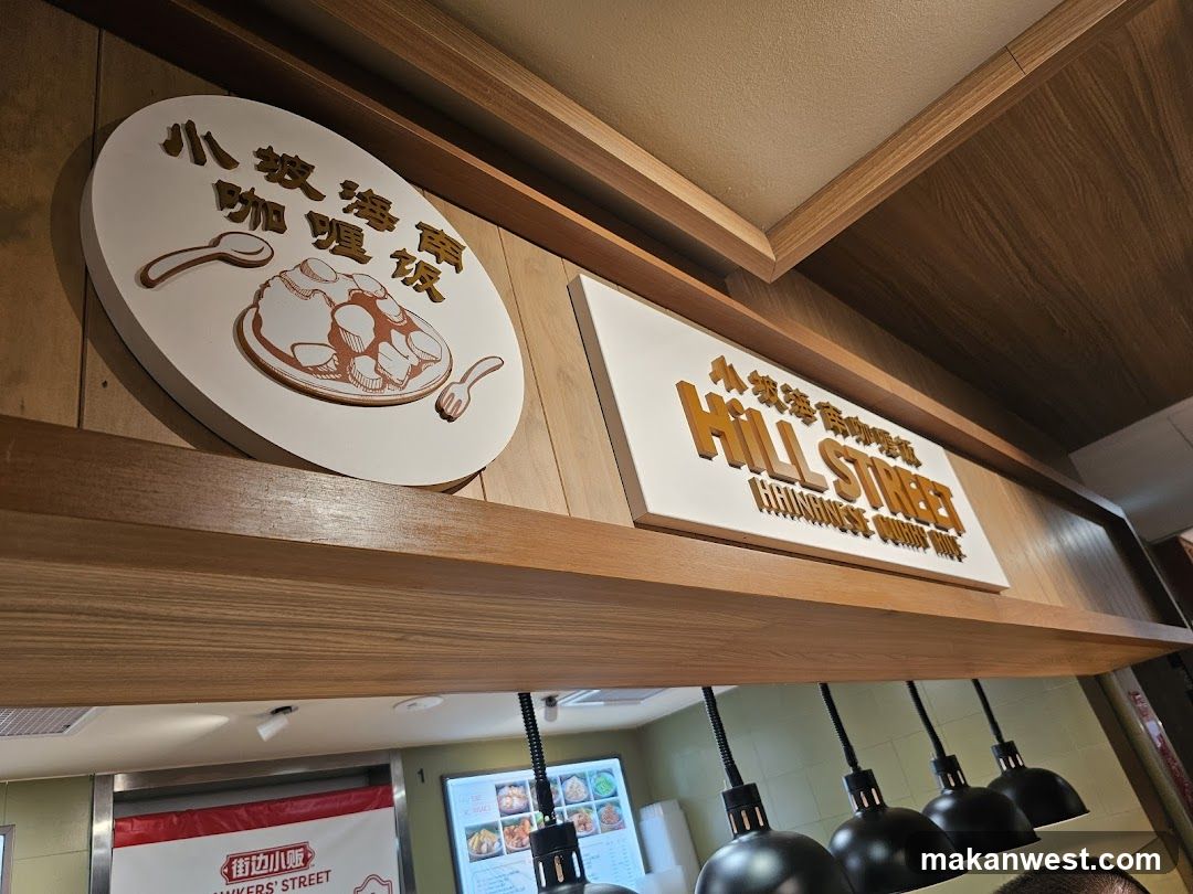 A close-up shot of signage for a restaurant. The main sign reads "HILL STREET HAINANESE CURRY RICE" in large, golden, 3D letters. Above this, in smaller golden Chinese characters, is more text. To the left, a circular sign depicts a bowl of food with a spoon and fork, also with Chinese characters above it. The signs are mounted on a wooden panel. Below the signs, a row of black heat lamps is visible, and further down, a digital menu board displays food items.