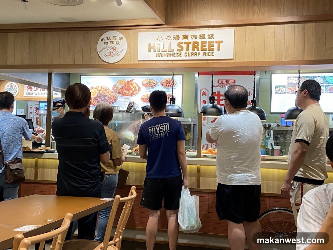 A group of people are standing in line at a food stall. The stall has a sign that says "HILL STREET HAINANESE CURRY RICE". There are pictures of food displayed above the counter.