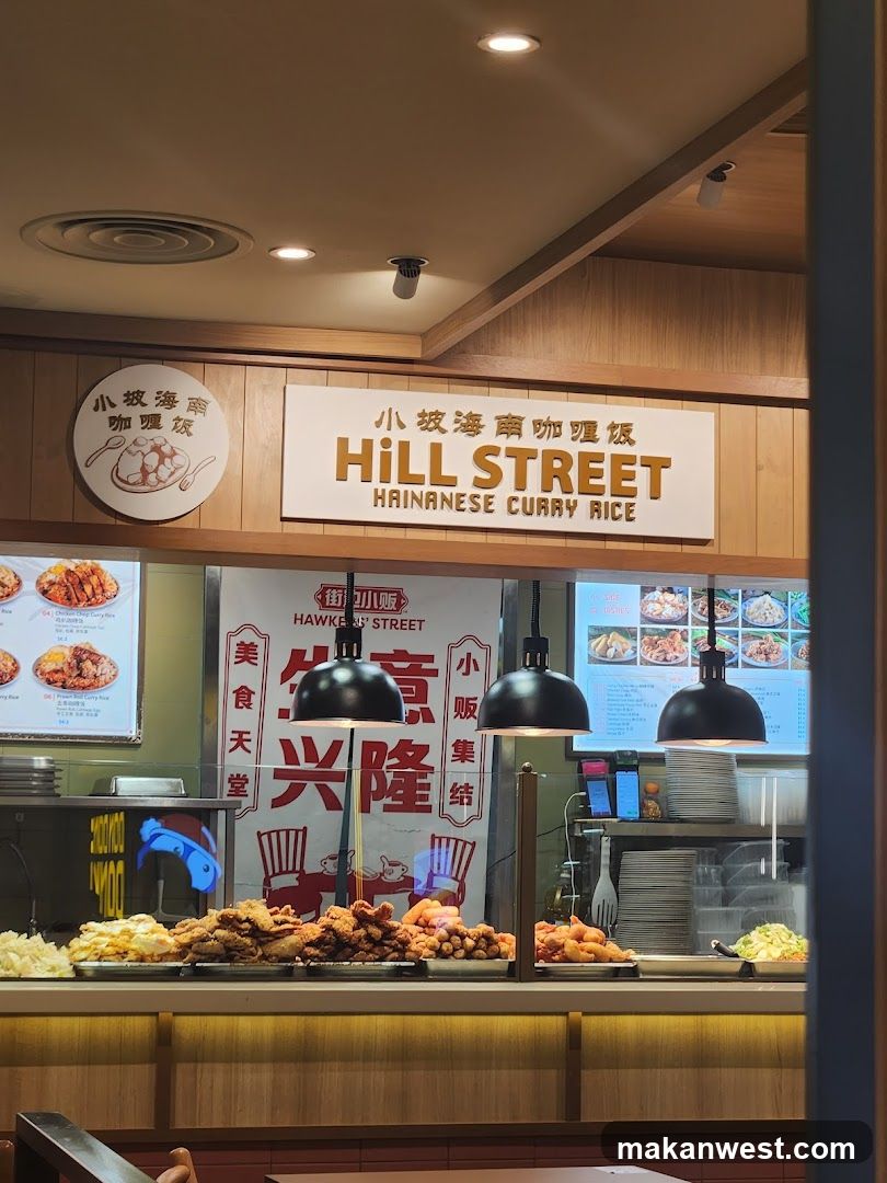 A food stall with a prominent sign reading "HILL STREET HAINANESE CURRY RICE" in English and Chinese. In front of the stall, various fried food items like chicken and sausages are displayed, along with some side dishes. A menu board with pictures of dishes is visible on the right. A red banner with Chinese characters and an illustration of chairs and a table is behind the counter.