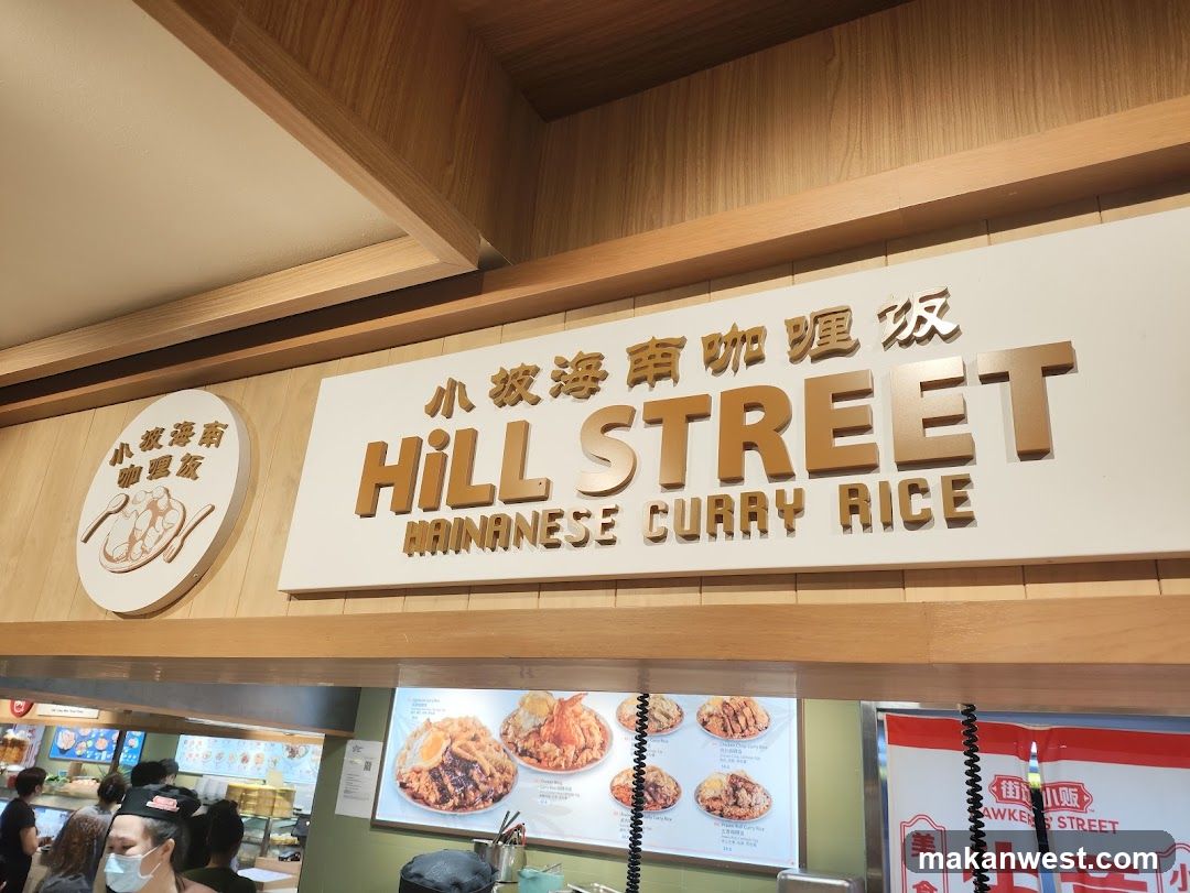 A sign for "Hill Street Hainanese Curry Rice" is prominently displayed. To the left, a circular sign features an illustration of a dish with spoons. Below the main sign, a menu board displays various curry rice dishes with images and prices. Several people are visible in the background, suggesting a food court or restaurant setting.