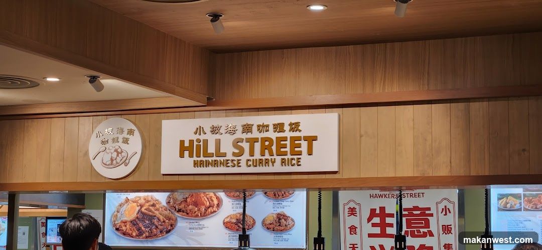 A sign for "Hill Street Hainanese Curry Rice" is displayed on a wooden paneled wall. Below the sign, a menu board shows various dishes with pictures and prices. A circular sign with Chinese characters and an illustration of a bowl of food is to the left of the main sign. The interior appears to be a food stall or restaurant.