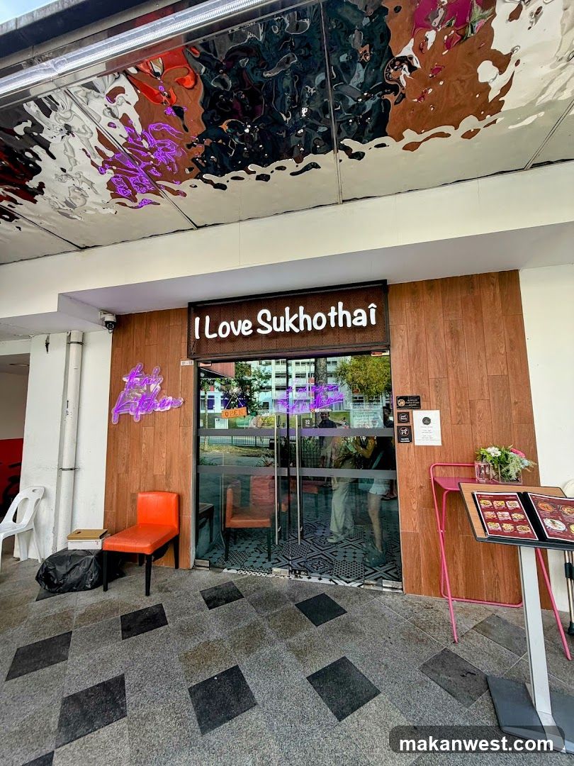 The storefront of a restaurant with a wooden facade and glass doors. A neon sign that says "I Love Sukhothai" is visible above the entrance. There are two orange chairs outside the entrance, one of which is occupied by a black trash bag. A menu stand with a pink frame is placed to the right of the entrance, displaying a menu with food pictures. The ceiling above the entrance is a reflective metallic surface with abstract patterns.