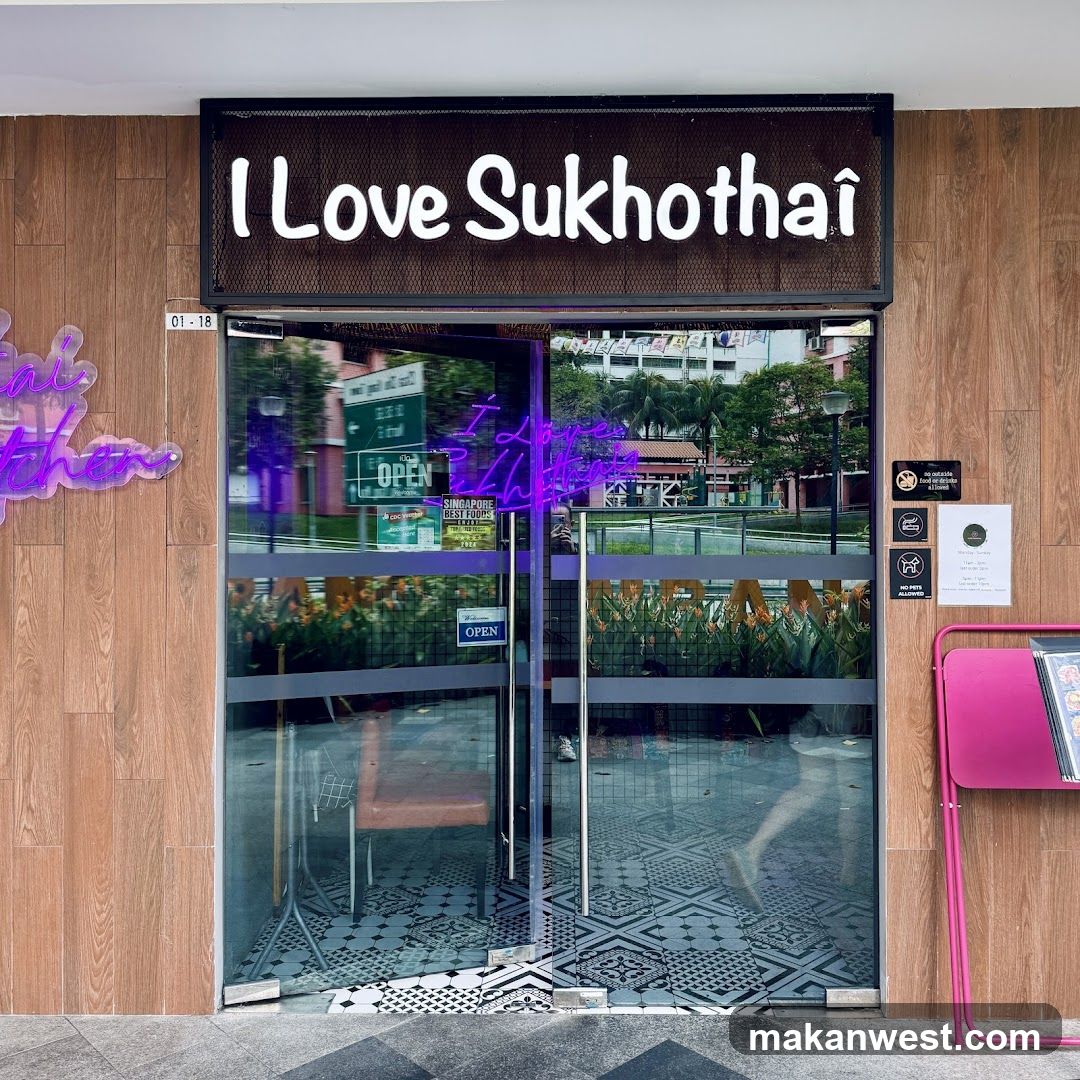 The storefront of a restaurant with a large neon sign that says "I Love Sukhothai". The entrance has glass doors with "OPEN" signs. There are also several smaller signs on the right side of the door. A pink stand with a menu is visible to the right of the entrance.