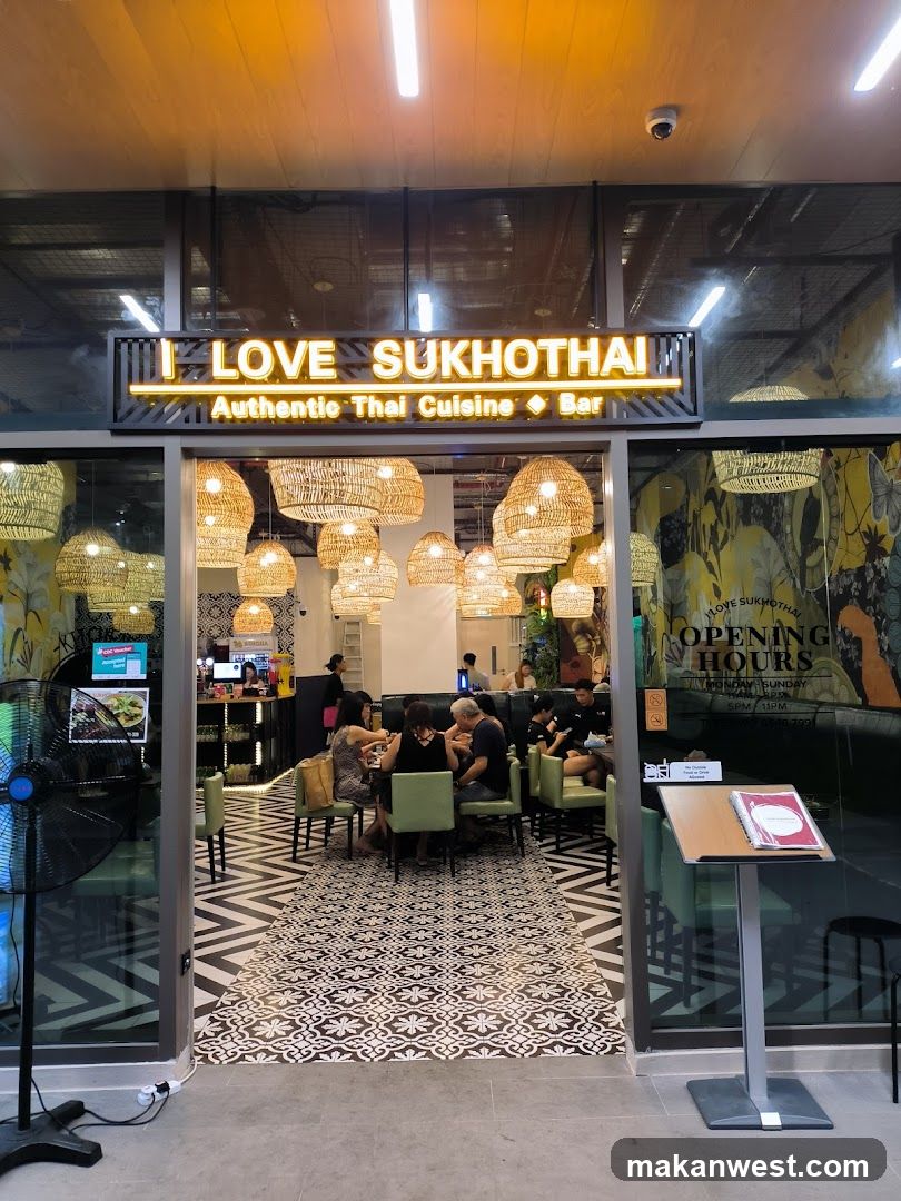 The exterior of a Thai restaurant with a prominent neon sign that reads "I LOVE SUKHOTHAI Authentic Thai Cuisine Bar". Large woven pendant lights hang from the ceiling. Inside, patrons are seated at tables, and the floor features a patterned tile design. A menu stand is visible near the entrance.