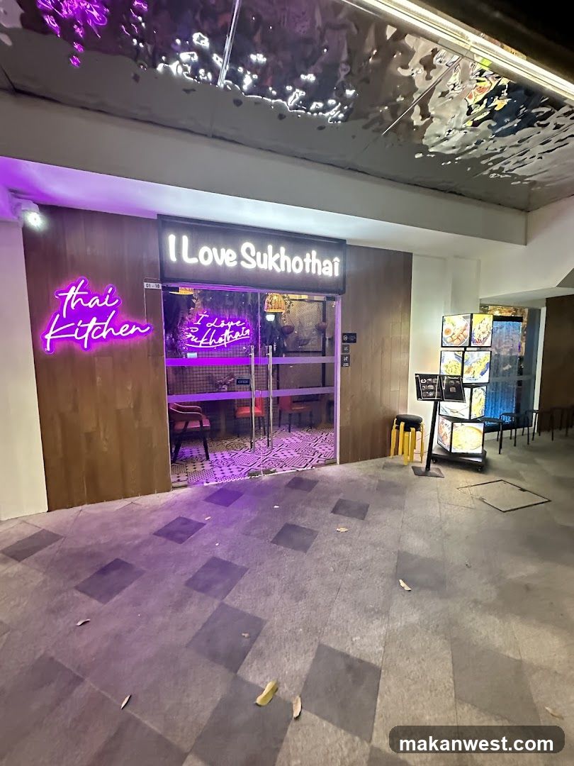 A storefront with neon signs reading "I Love Sukhothai" and "thai kitchen". The entrance has glass doors revealing a tiled floor and chairs inside. A menu display stand is visible to the right.