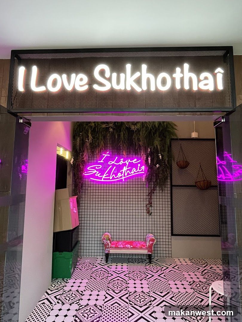 A storefront with a large illuminated sign that reads "I Love Sukhothai". Below the sign, a neon sign in pink also reads "I Love Sukhothai". There is a small decorative bench with a floral pattern in front of a black grid wall. Hanging plants are above the bench. The floor is covered in a black and white patterned tile.