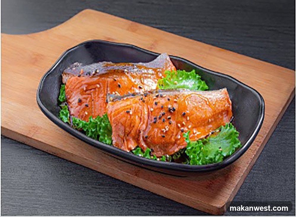 Two pieces of glazed salmon are served on a bed of lettuce in a black oval dish. The salmon is topped with sesame seeds. The dish is placed on a wooden board.