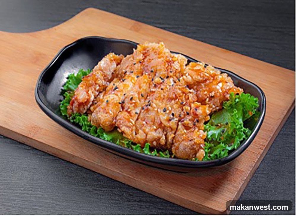 A close-up shot of crispy fried chicken pieces, coated in a glossy sauce and sprinkled with black and white sesame seeds. The chicken is served on a bed of fresh green lettuce leaves in a black, irregular-shaped bowl. The bowl rests on a wooden board, which is placed on a dark textured surface.