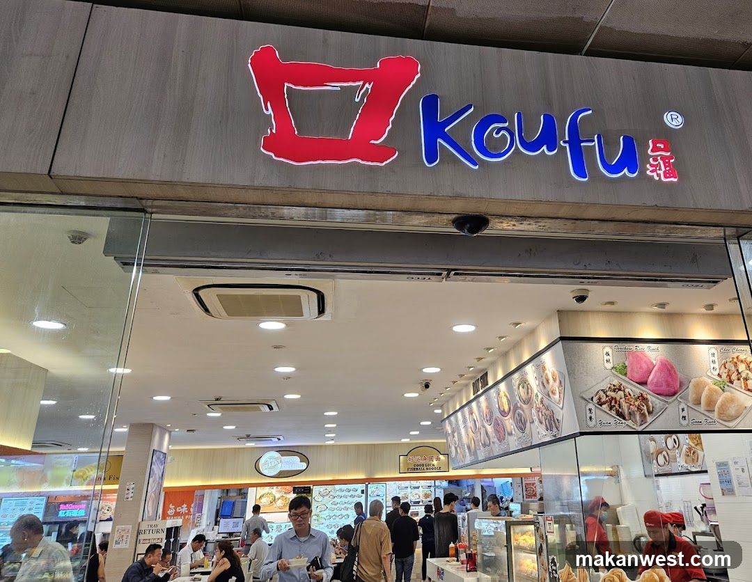 The exterior of a Koufu restaurant with its prominent red and blue signage. Inside, customers are seen dining and ordering food. Displayed on the walls are images of various dishes, including "Guan Yang", "Siu Kock", and "Peachbun Rice Bunch".