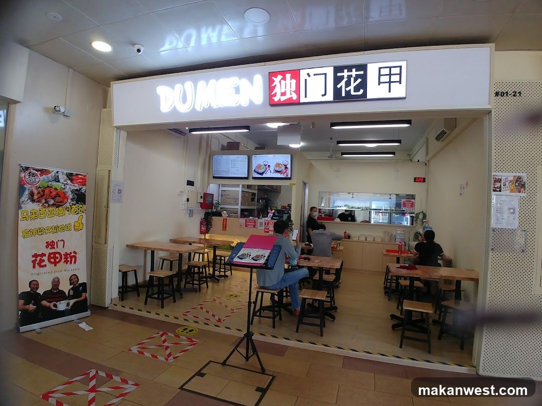 The exterior of a restaurant with a prominent illuminated sign that reads "DUMEN" and Chinese characters. Inside, tables and chairs are set up for dining. A large banner with images of food and text in Chinese and English is displayed to the left of the entrance. The restaurant appears to be open for business.