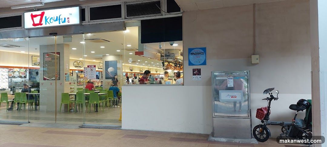 The exterior of a Koufu food court with a large glass storefront. Inside, green tables and chairs are visible, along with customers dining and staff working behind a counter. A prominent illuminated sign with the Koufu logo is displayed above the entrance. To the right of the entrance, there is a stainless steel collection box and a parked electric bicycle.