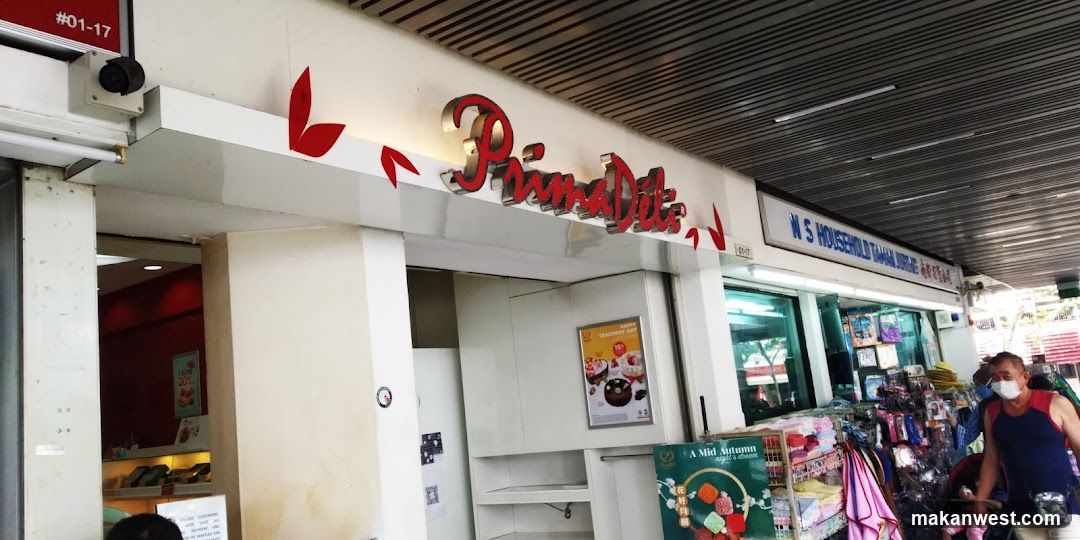 The exterior of a PrimaDeli shop with its prominent red and silver logo above the entrance. To the right, a sign reads "N S HOUSEHOLD TAMAN JURONG". The shop displays various items for sale, including what appear to be baked goods and household products. A poster for "Happy Teacher's Day" with a 15% discount is visible.