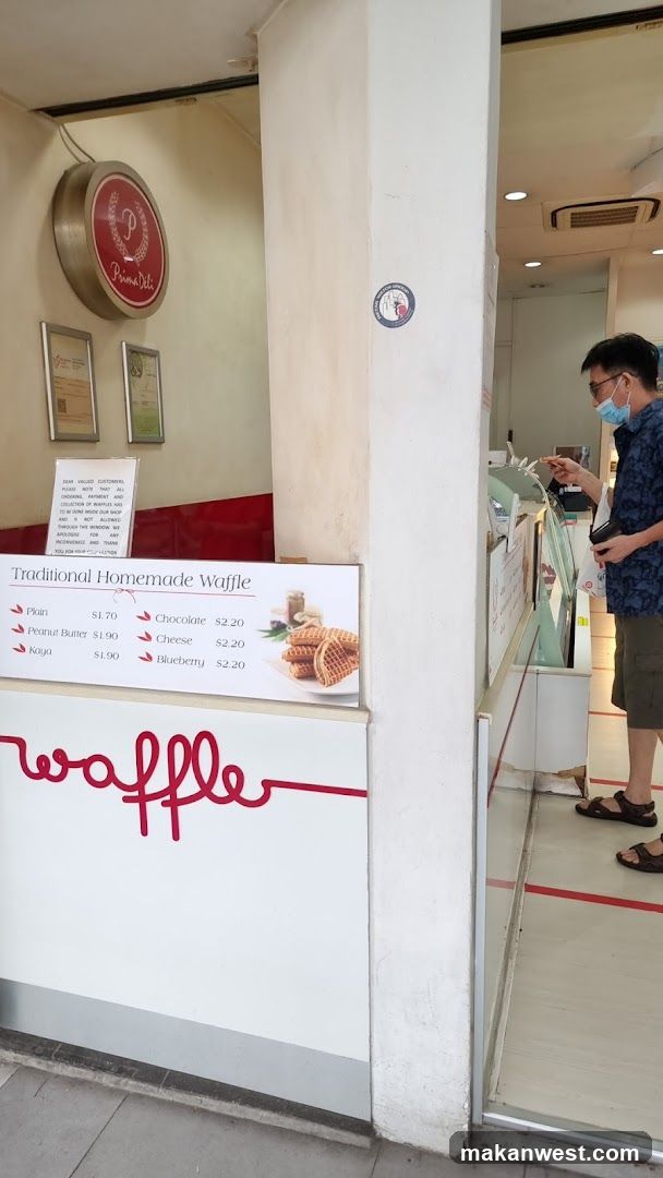 A man wearing a mask stands at a counter inside a shop. A sign on the wall reads "Prima Deli" and a menu board lists traditional homemade waffles with various toppings. The word "waffle" is written in large red script on the counter.