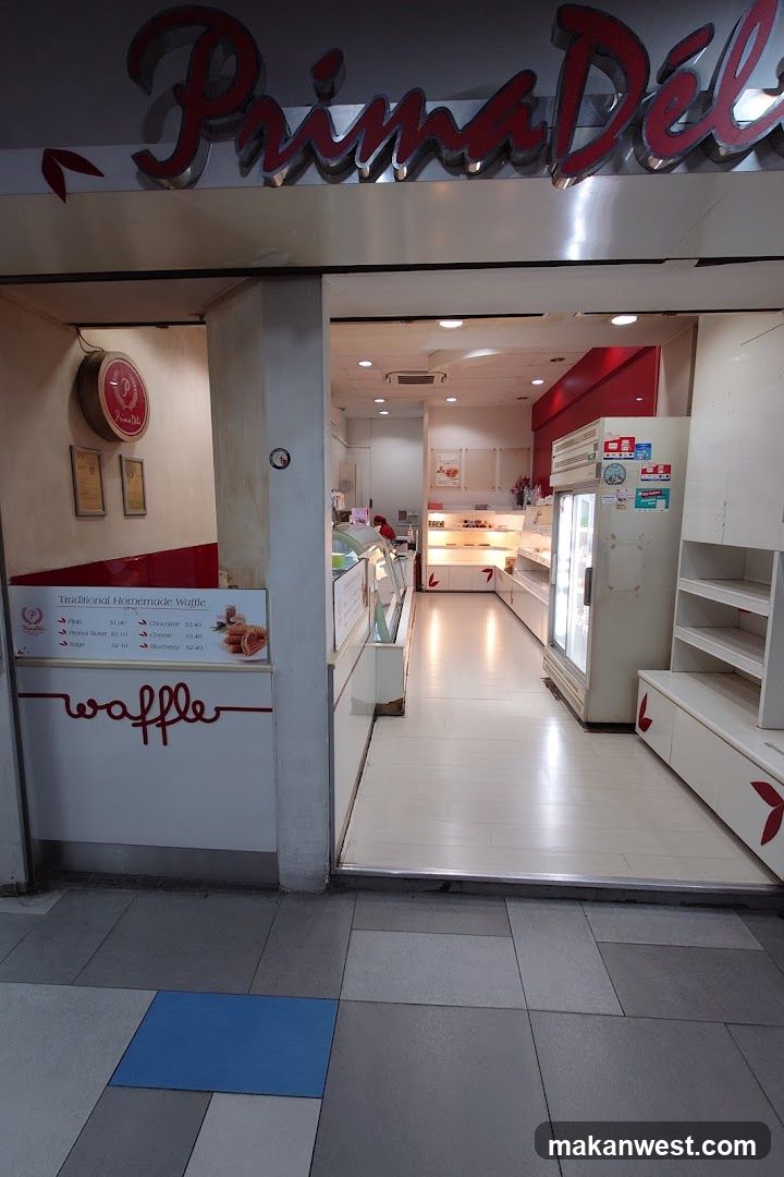 The exterior of a Prima Deli shop. The shop has a large red and silver sign above the entrance. Inside, the shop is brightly lit and appears to be a bakery or dessert shop with display cases and shelves. A menu board is visible to the left of the entrance.