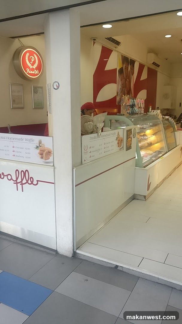 A view of the Prima Deli shop front with a circular sign featuring the logo and name. There are framed certificates on the wall. A display counter with a glass case filled with pastries is visible, along with a menu board advertising traditional homemade waffles. A person is seen behind the counter.