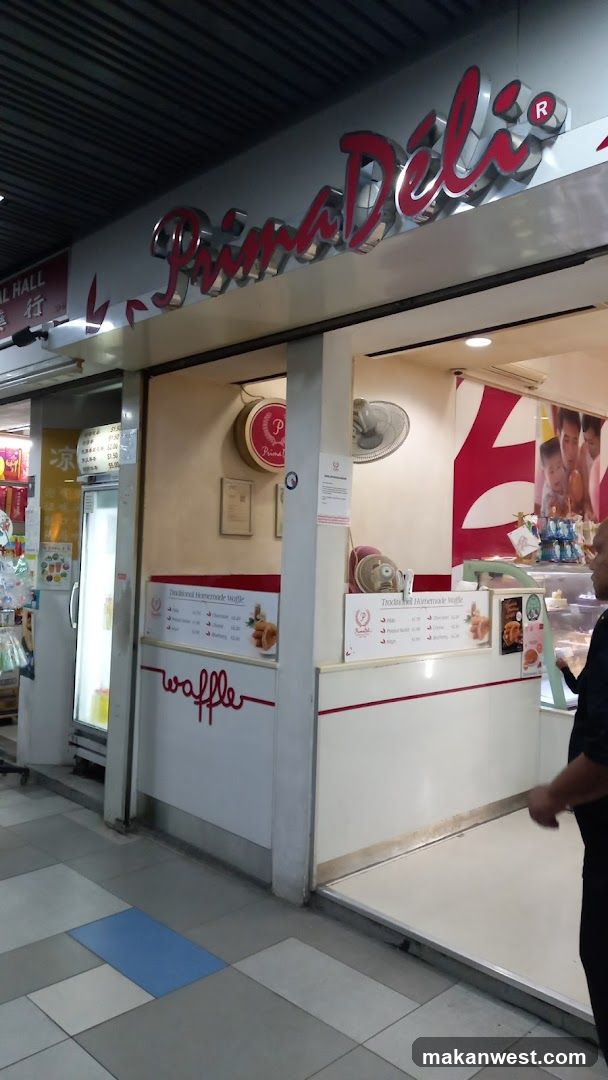 The exterior of a Prima Deli shop. A large sign with the shop's name is visible above the entrance. Inside, there are display cases and a counter. A menu board is visible on the counter.
