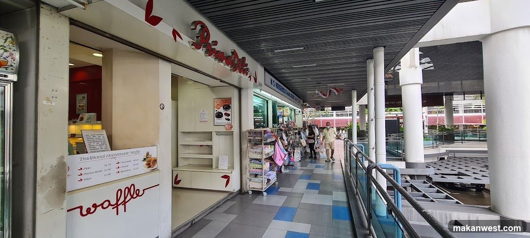 The exterior of a PrimaDeli shop with a prominent red sign. To the left, a waffle stand displays a menu with various waffle options and prices. The shop interior is visible, showing shelves and a red-colored wall. A rack of merchandise is placed outside the shop.