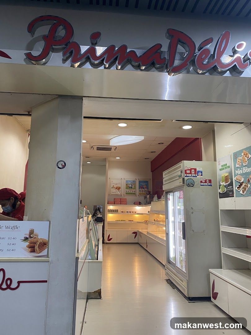 The exterior of a bakery with a prominent sign reading "Prima Déli". Inside, display cases show pastries and baked goods. A refrigerator is visible, along with shelves stocked with products. A menu board displays waffle prices.