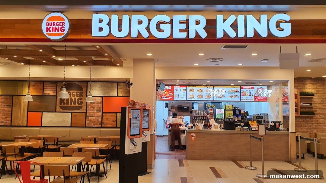 The exterior of a Burger King restaurant with its prominent signage. The interior shows a counter area with staff, menu boards displaying food items, and seating areas with tables and chairs. Digital ordering kiosks are also visible near the entrance.