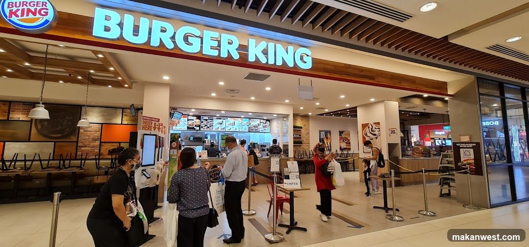 The exterior of a Burger King restaurant with its prominent signage. Inside, customers are seen ordering at the counter and using self-service kiosks. The dining area has tables and chairs, some of which are stacked. The overall ambiance is that of a fast-food establishment within a mall or shopping center.