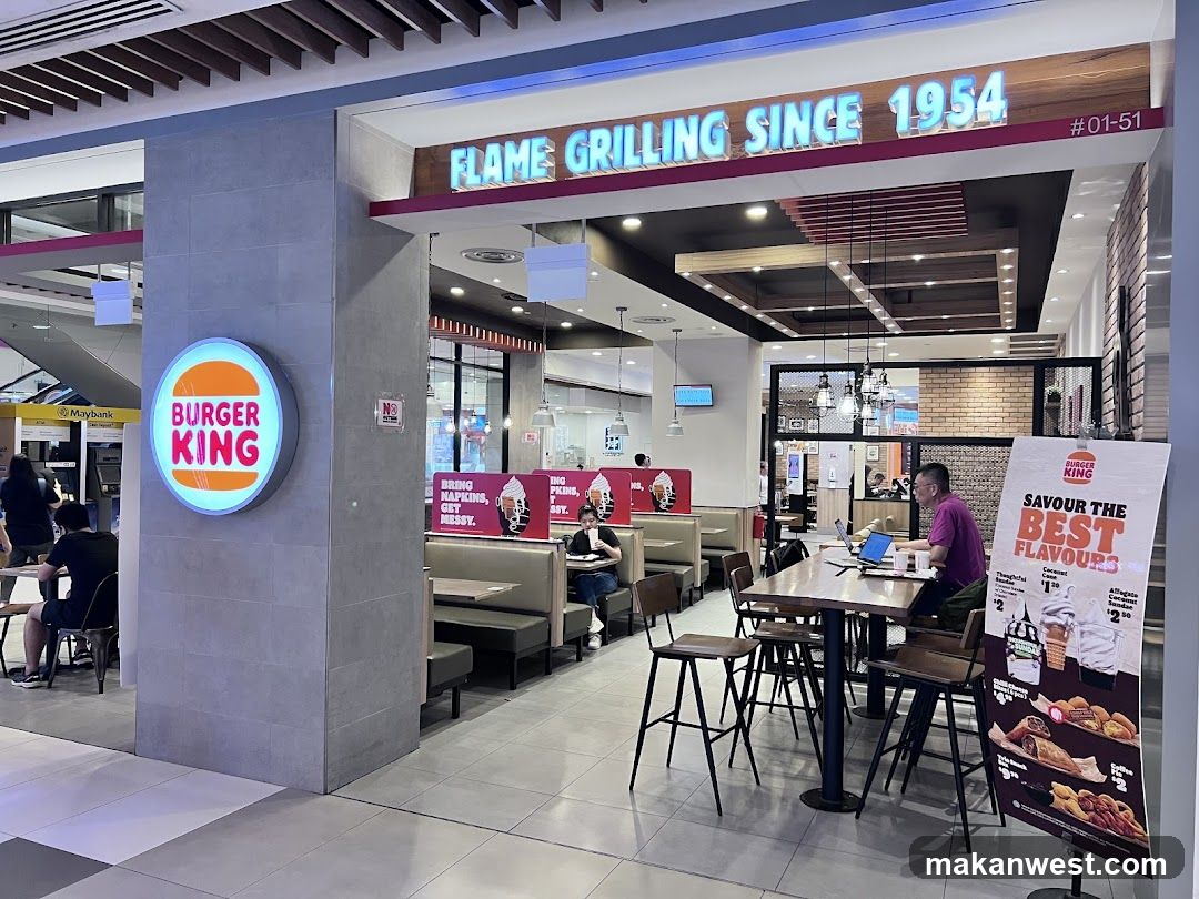 The exterior of a Burger King restaurant with its signature circular logo and "Flame Grilling Since 1954" signage visible. Inside, booths and tables are set up, with some customers seated. A promotional poster is displayed near the entrance.