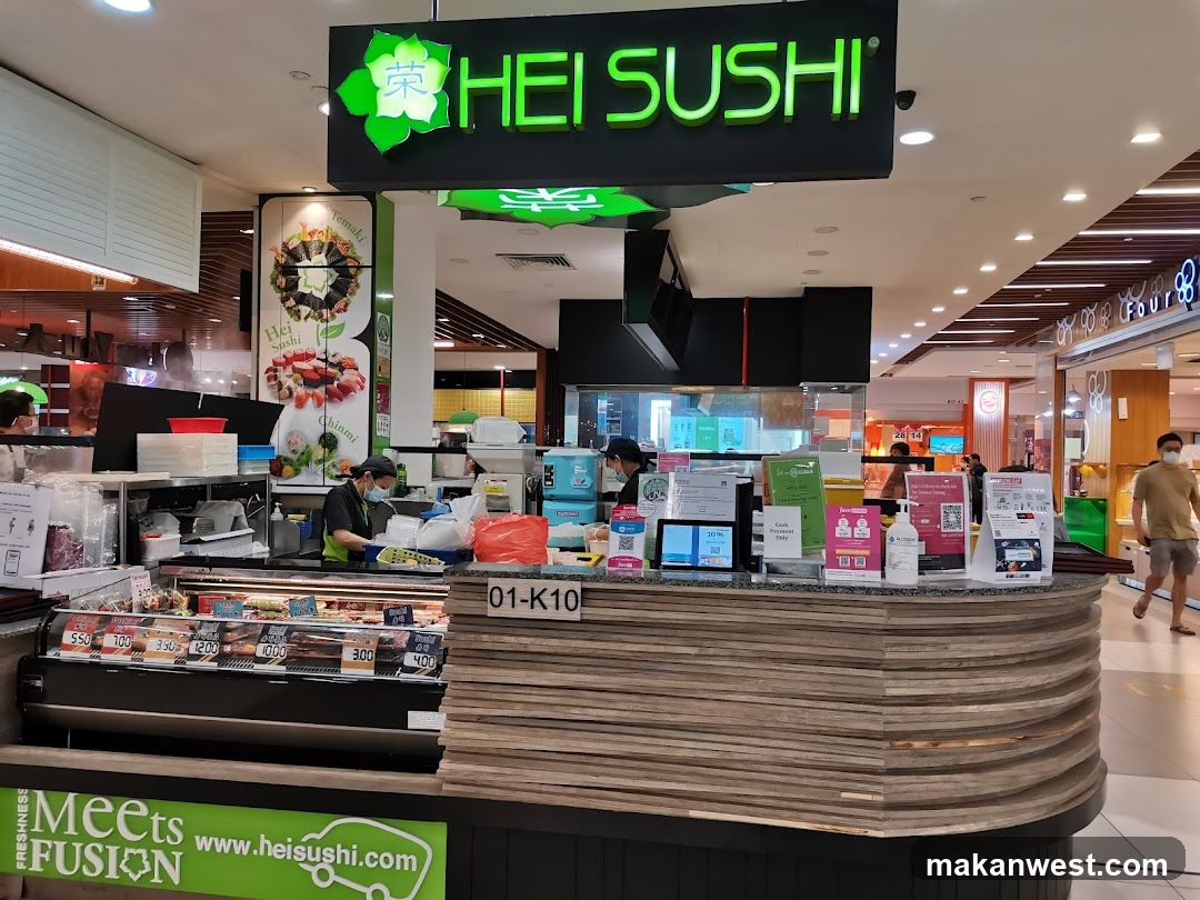 A sushi stall with a prominent "HEI SUSHI" sign. The stall displays various sushi items in a refrigerated case with prices. Staff members are visible behind the counter preparing food. The stall is located within a shopping mall.