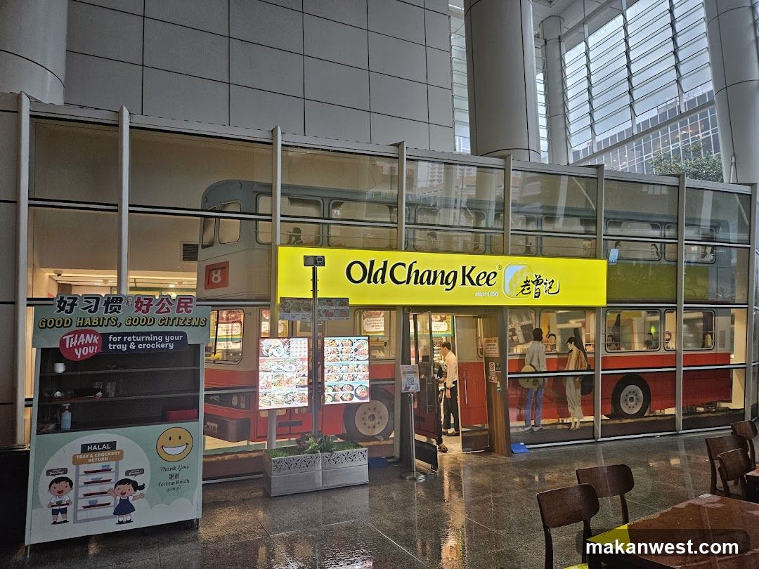 The exterior of an Old Chang Kee restaurant designed to look like a double-decker bus. A yellow sign with the restaurant's name is prominently displayed. There are menu boards visible, and people can be seen inside the establishment. A separate stand with the text "GOOD HABITS, GOOD CITIZENS" and "HALAL TRAY & CROCKERY" is also present.