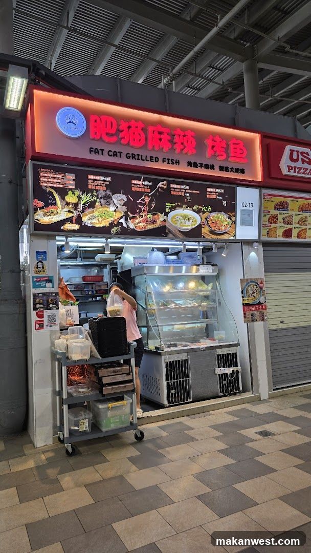 A food stall with a prominent sign that reads "FAT CAT GRILLED FISH" in English and Chinese. The stall displays various grilled fish dishes and other food items in a refrigerated display case. A person is seen preparing food behind the counter. Adjacent to the stall is a "US PIZZA" stall.
