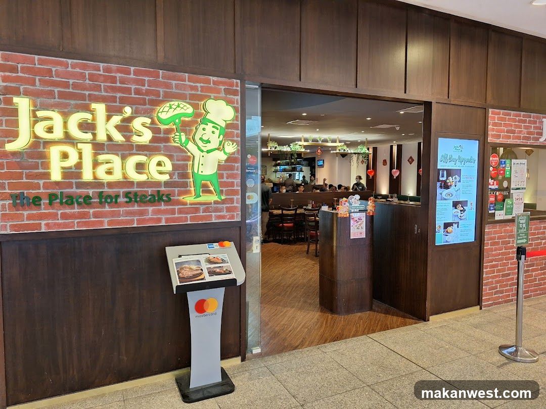 The exterior of a restaurant called Jack's Place, which advertises itself as "The Place for Steaks." The entrance is open, revealing a dining area with tables and patrons. A digital menu board is visible to the right of the entrance, displaying food items and promotions. A payment terminal with Mastercard branding is located to the left of the entrance.