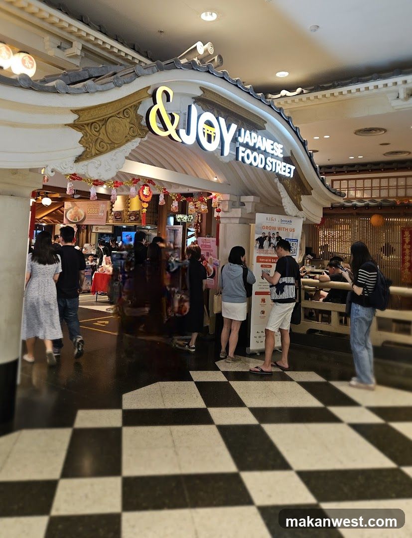 The exterior of a Japanese food street establishment with a prominent sign that reads "&JOY JAPANESE FOOD STREET". The entrance is decorated with traditional Japanese architectural elements and festive red lanterns. Several people are seen entering and standing near the entrance. The floor has a black and white checkered pattern.