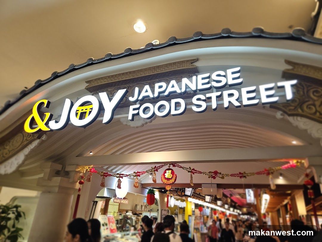 A brightly lit sign above an archway reads "& JOY JAPANESE FOOD STREET". Below the sign, a bustling food court is visible with many people walking around and stalls lining the sides. Festive red and gold decorations hang from the archway.