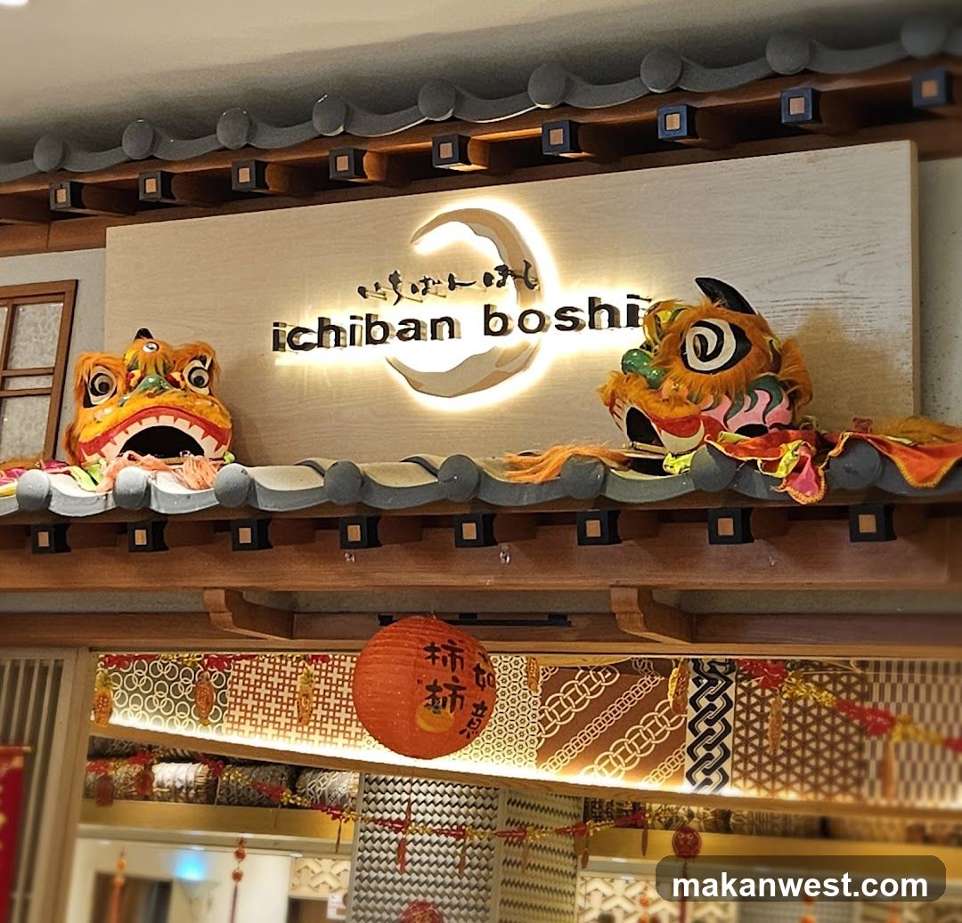 A brightly lit sign with the words "ichiban boshi" in English and Japanese characters is mounted on a light-colored panel. The sign is illuminated from behind and features a crescent moon graphic. Two decorative lion heads, one on each side, are placed on a tiled roof structure above the sign. Below the sign, an orange lantern with Chinese characters hangs, and festive decorations with traditional patterns are visible.