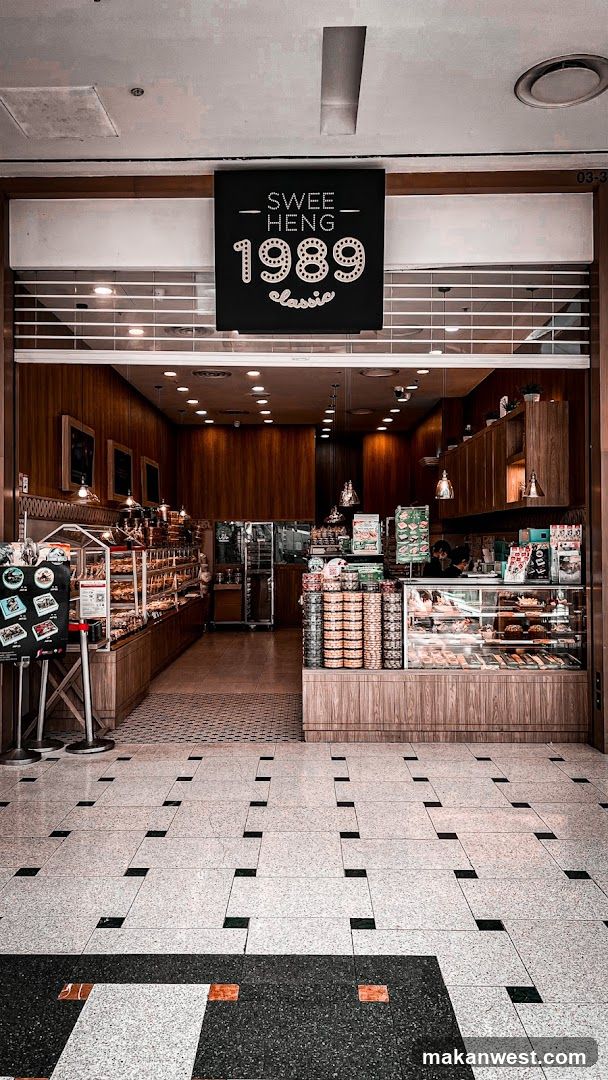The exterior of a bakery with a prominent black sign displaying "SWEE HENG 1989 classic" in white lettering. The entrance is wide, revealing a well-lit interior filled with display cases of baked goods and shelves stocked with packaged items. The floor is a patterned tile mosaic.