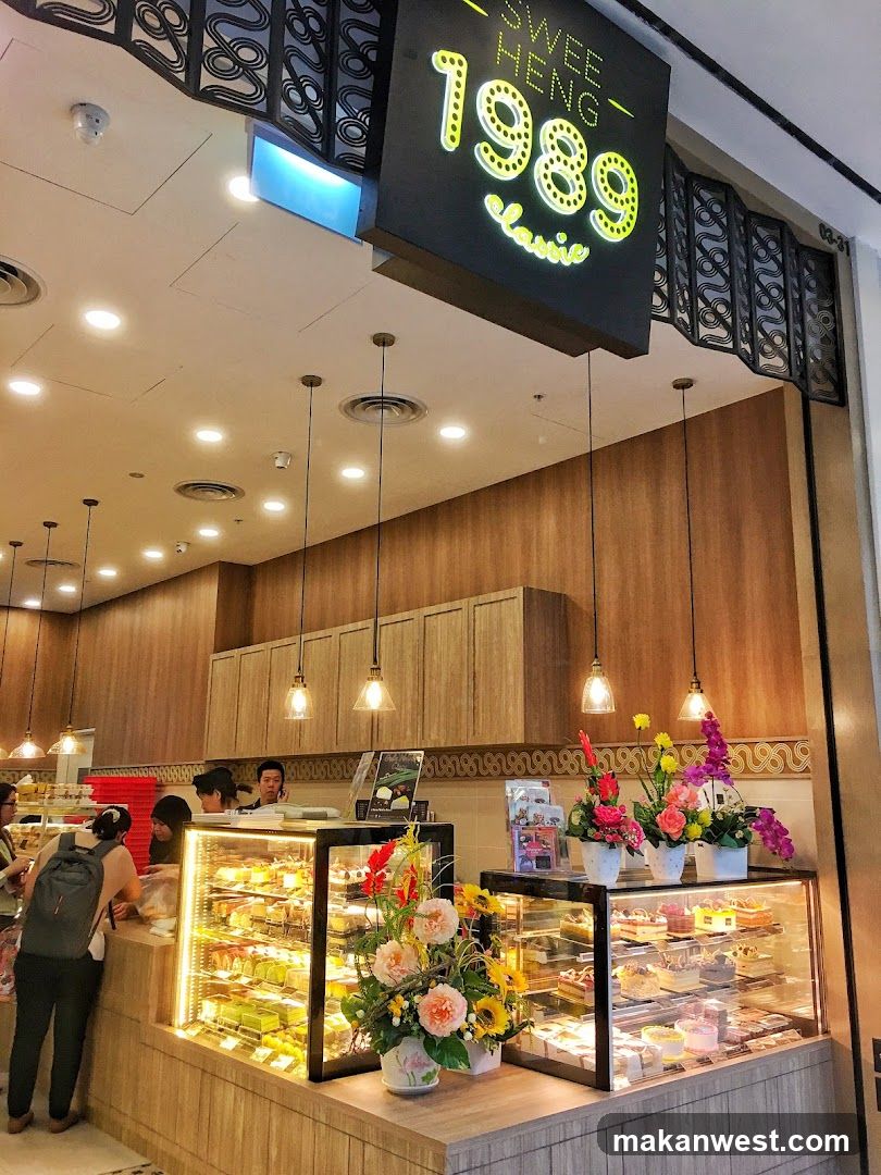 A view of the interior of a bakery with a prominent sign that reads "SWE HENG 1989 Classic". The bakery features display cases filled with various cakes and pastries. Customers are seen interacting with staff at the counter. The overall ambiance is bright and inviting, with wooden accents and decorative floral arrangements.