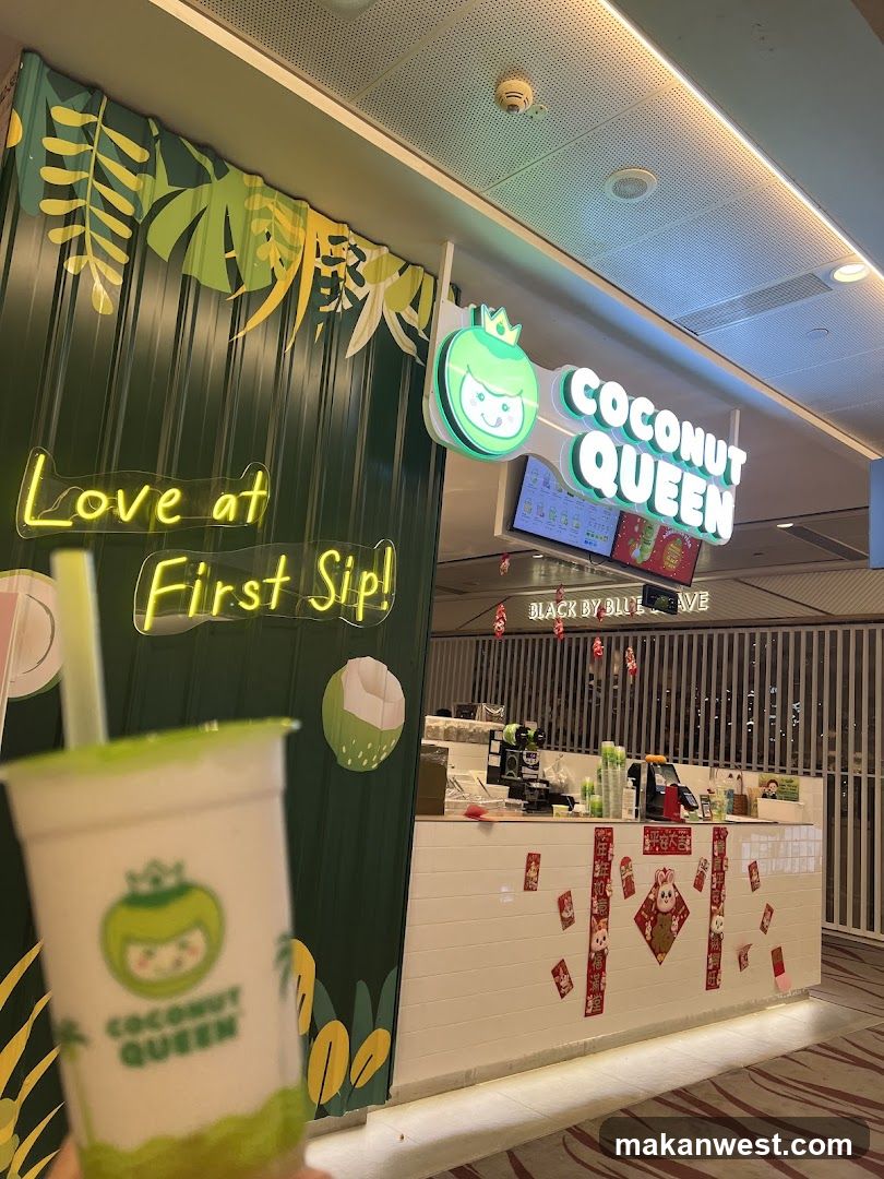 A bright and inviting storefront for Coconut Queen, a beverage shop. The facade features a vibrant green corrugated metal wall adorned with tropical leaf cutouts and a neon sign that reads "Love at First Sip!". Above, a prominent illuminated sign displays the shop's name, "COCONUT QUEEN", alongside a cheerful cartoon coconut character wearing a crown. The counter area is decorated with festive red and gold Chinese New Year ornaments, including rabbit imagery. A hand holds a Coconut Queen branded drink in the foreground.